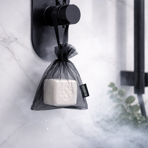 Hanging Silknsoak shower steamer cube in organza bag, releasing eucalyptus aroma steam in a modern white tile shower.